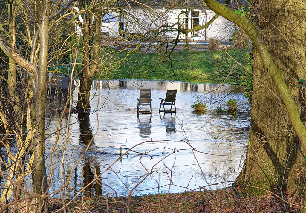 Blick durch kahle Äste auf eine überschwemmte Gartenfläche am Osteufer. Zwei hölzerne Gartenstühle stehen mitten im Hochwasser, im Hintergrund ist ein weißes Wohnhaus zu sehen.
