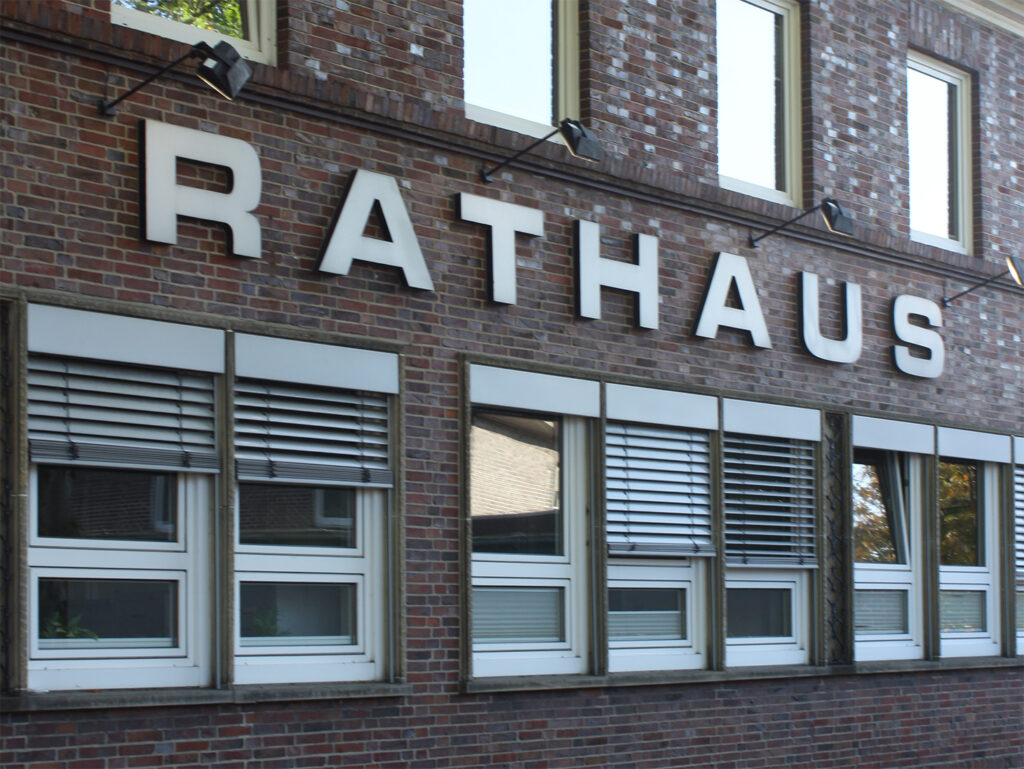 Die Fassade eines dunklen Backsteingebäudes mit einem großen, weißen Schriftzug "RATHAUS" über den Fenstern des Erdgeschosses. Die Fenster im unteren Bereich haben Außenjalousien, die teilweise geschlossen sind. Oberhalb des Schriftzugs sind mehrere schwarze Lampen angebracht die den Schriftzug anleuchten.